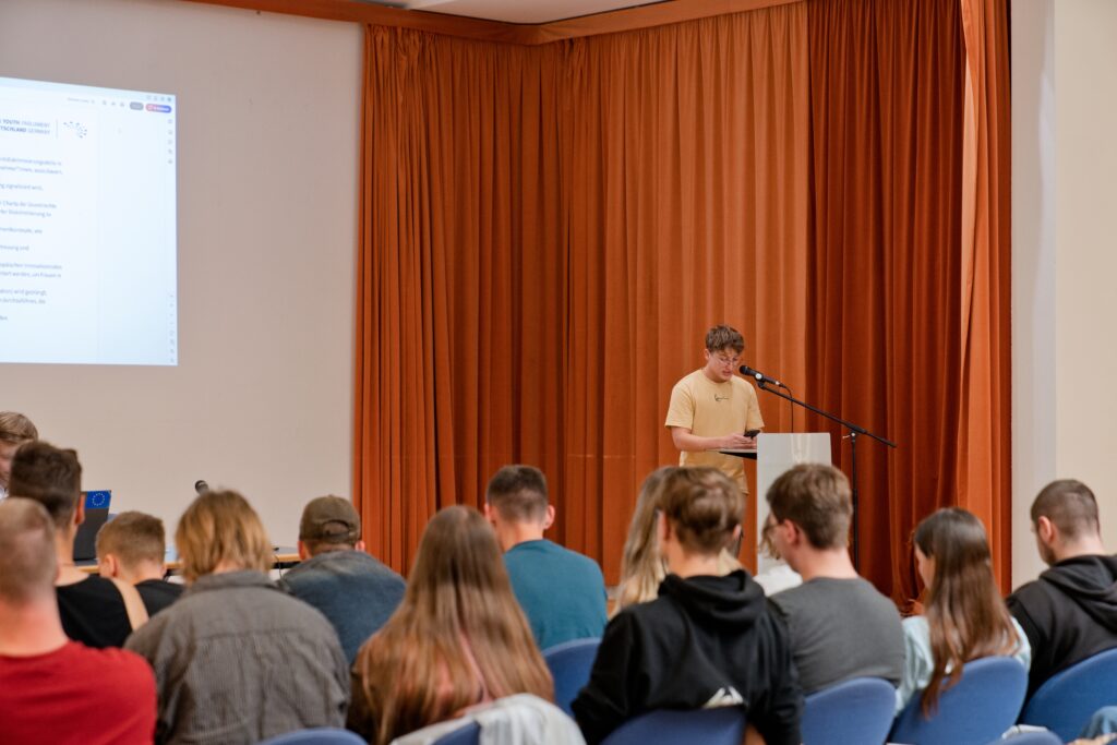Das Bild zeigt im Foksu einen Person an einem Podium. Diese Person hält eine Rede. Im vorderen Teil des Bildes sieht man die Hinterköfpe einiger weiterer Personen, die der Rede zuhören.